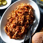Pulled chicken on a plate with pickles and buns.