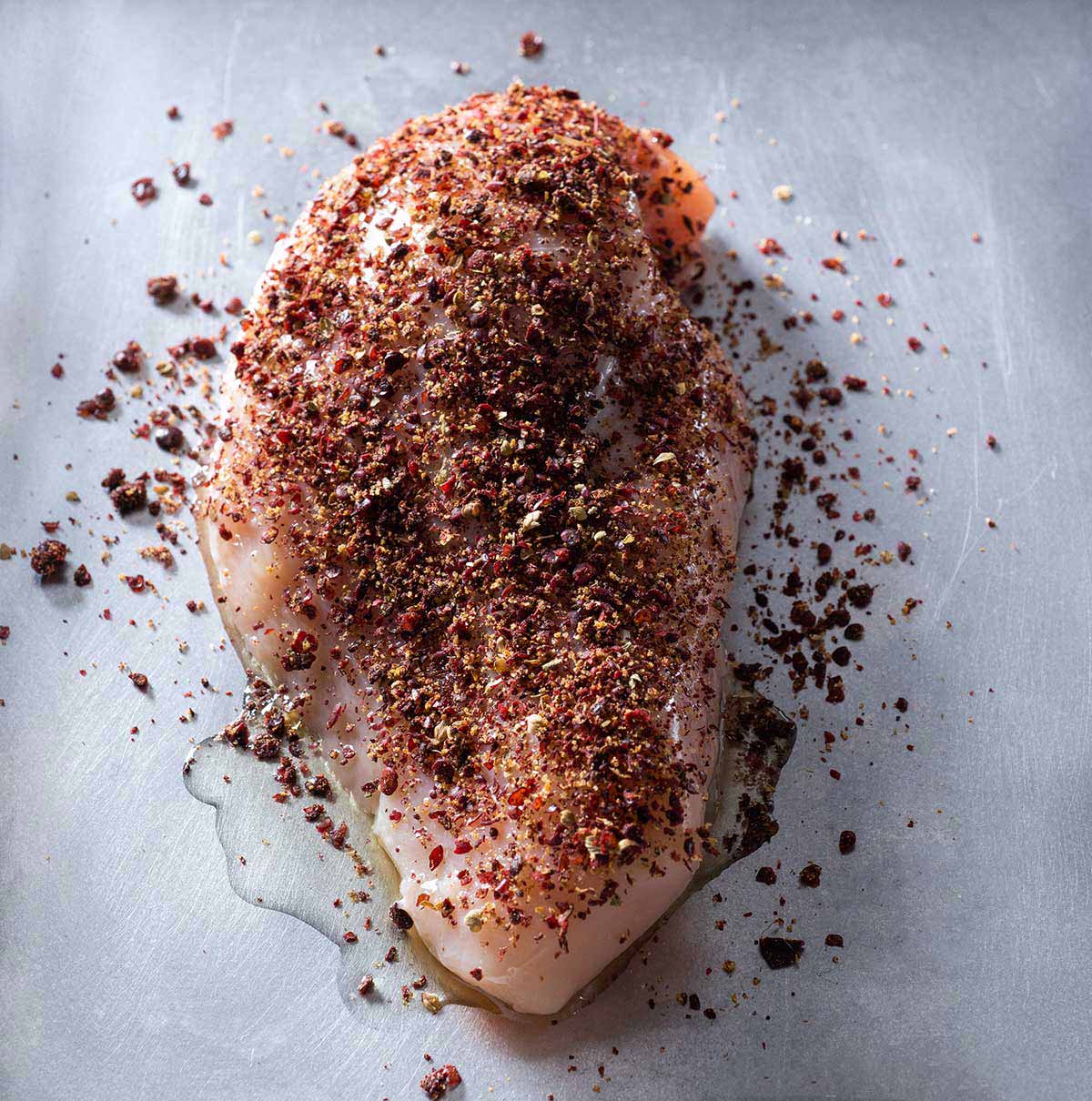 A raw chicken breast with sumac dry rub on a baking sheet.